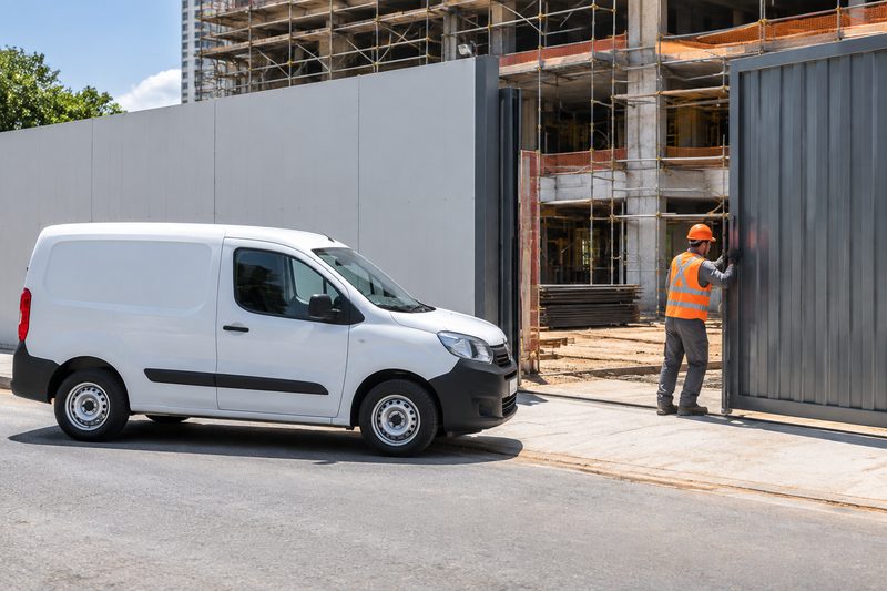 Van de carga branca esperando na rua enquanto um trabalhador com capacete abre o portão de uma obra