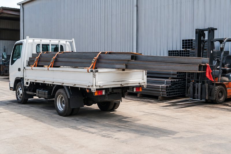 Caminhão de carroceria aberta com vigas metálicas compridas amarradas com cintas e sinalizadas na extremidade