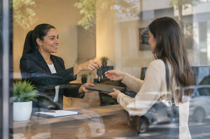 Atendente entregando a chave e uma pasta a um cliente em uma locadora de veículos, visto através da vidraça