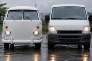Frente arredondada de uma Kombi de carga branca ao lado da frente quadrada de uma van branca, com chuva leve e asfalto molhado